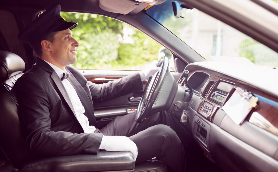 Chauffeur in a dark suit and cap seated behind the wheel of a luxury vehicle