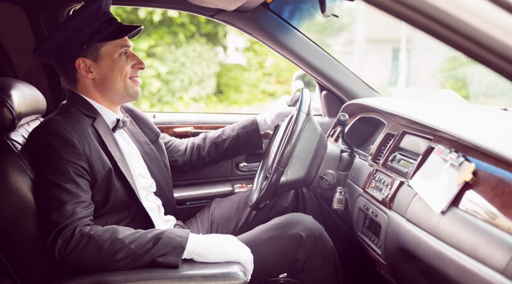 Chauffeur in a dark suit and cap seated behind the wheel of a luxury vehicle